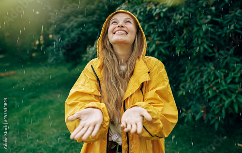 Image of a positive young blonde woman smiling wearing yellow raincoat during the rain in the park. Cheerful female enjoying the rain outdoors. Woman looking up and catching the rain drop with hands. photo