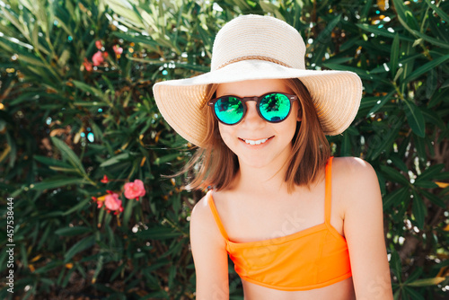 Little smiling playful girl in a straw hat and sunglasses - looking directly at the camera - against the background of a bush with flowers - carefree concept