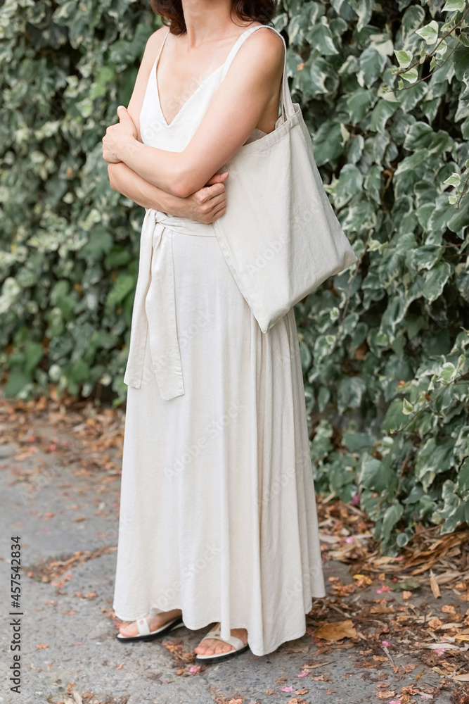 Linen reusable bag on a shoulder of a girl in a natural dress against the background of a green wall of leaves. Eco-friendly product concept. Linen reusable bag on a shoulder of a girl in a natural dress against the background of a green wall of leaves. Eco-friendly product concept.