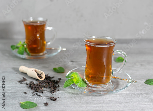 Two Turkish cups of tea stand on a light wooden table. Mint leaves and tea leaves are scattered in the background