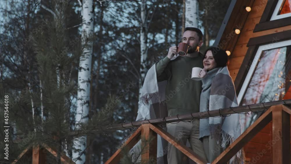 PAN slowmo of couple wrapped in warm blanket drinking hot tea on porch of A-frame cabin in woods on cold evening PAN slowmo of couple wrapped in warm blanket drinking hot tea on porch of A-frame cabin in woods on cold evening
