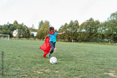 little boy child in a superhero costume is playing football in the fresh air. happy boy in a red raincoat and a mask runs with a ball on the football field. Outdoor games, dynamic image