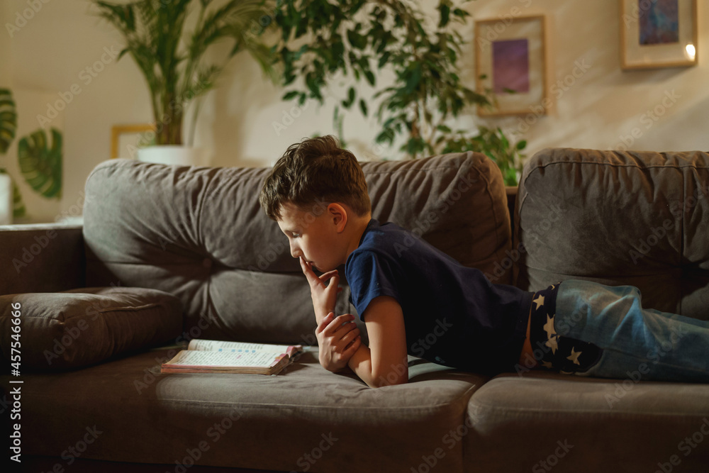 An attentive boy is reading a book lying on the couch at home. The student is passionate about reading. The child prepares homework, does homework lying down.