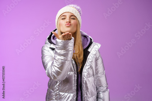 Romantic tender gentle cute girlfriend blond hair wearing hat stylish silver jacket close eyes folding lips muah hold hand near lips blow air kiss passionately feel love sympathy, purple background photo