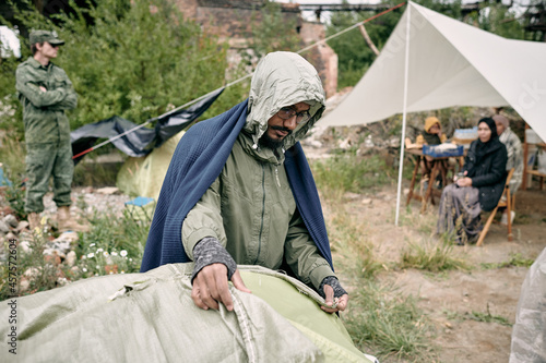 Sorrowful Arabian refugee man with blanket on shoulders covering tent with sack while preparing for cold weather, solder in background