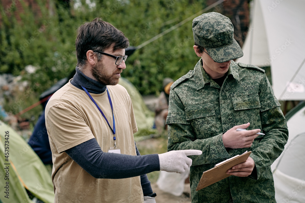 Soldier in camouflage wear holding clipboard and pointing aside while discussing food supply with social worker in migrant camp