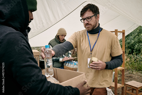 Young bearded volunteer in glasses holding tin can and giving bottle of water to homeless man with box photo