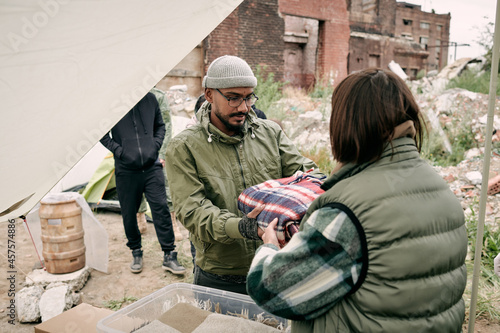 Rear view of social worker in warm vest giving plaid to middle-eastern migrant in glasses and hat outdoors photo