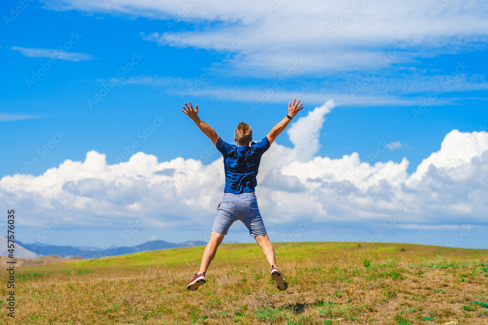 Rear view of a happy young man jumping on a summer meadow in the air against a blue sky with clouds Rear view of a happy young man jumping on a summer meadow in the air against a blue sky with clouds