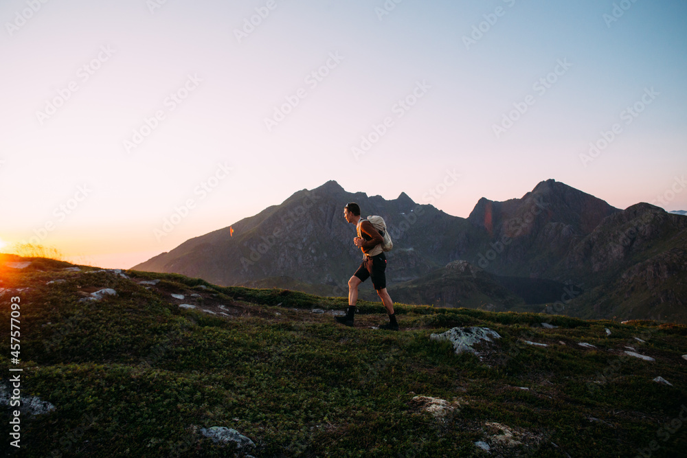 Fototapeta premium Young athletic man with backpack hike to reach summit of mountain at sunset. Cinematic beautiful adventure. Inspiring time spent outdoors. Explore norway in summer