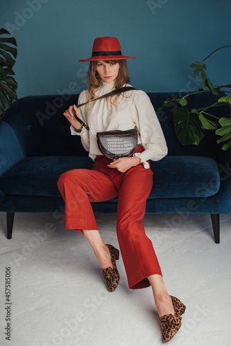 Autumn fashion conception: elegant  woman wearing stylish orange hat, wide culottes, white silk blouse, leopard print loafer shoes, with brown faux leather sholder bag, sitting on blue sofa.  photo