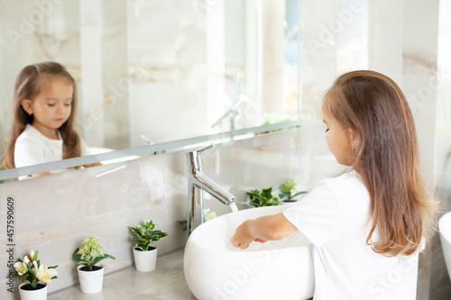 A beautiful cute little girl washes her hands in the bright bathroom in front of the mirror. Hygiene and cleanliness. Prophylaxis. Beautiful interior photo