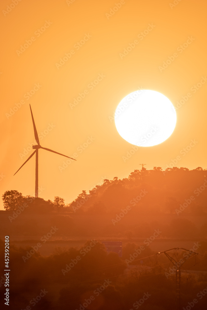 Vertical image of Sunrise with a large sun rising from behind a mountain next to a renewable energy wind turbine during the golden hour. Vertical image of Sunrise with a large sun rising from behind a mountain next to a renewable energy wind turbine during the golden hour.