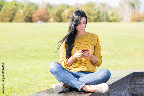 A beautiful and attractive Caucasian brunette girl sitting and using a smartphone in the park.