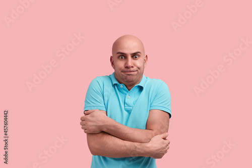 Im trembles. Scared bald homosexual man with bristle feels cold, hugs himself to warm up, clatter with teeth, gay friendly, wears blue polo shirt, poses over pink background.