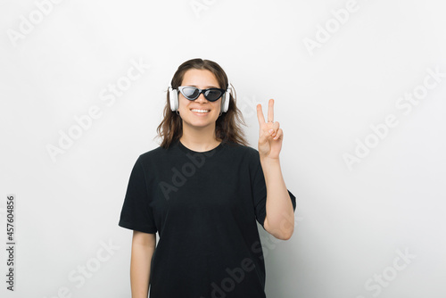 Stylish female student wearing headphones is showing the v or peace gesture over white background.