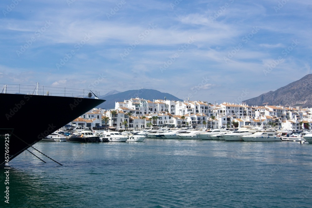The foreground of a larger yacht is visible in the foreground. In the background you can see the port of Puerto Banús and the mountains around Marbella.