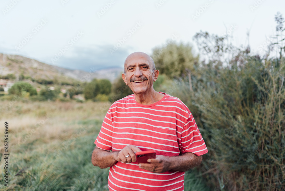 Middle-aged man looking at the camera and smiling while using a mobile phone outdoors in the countryside. Technology concept. Middle-aged man looking at the camera and smiling while using a mobile phone outdoors in the countryside. Technology concept.
