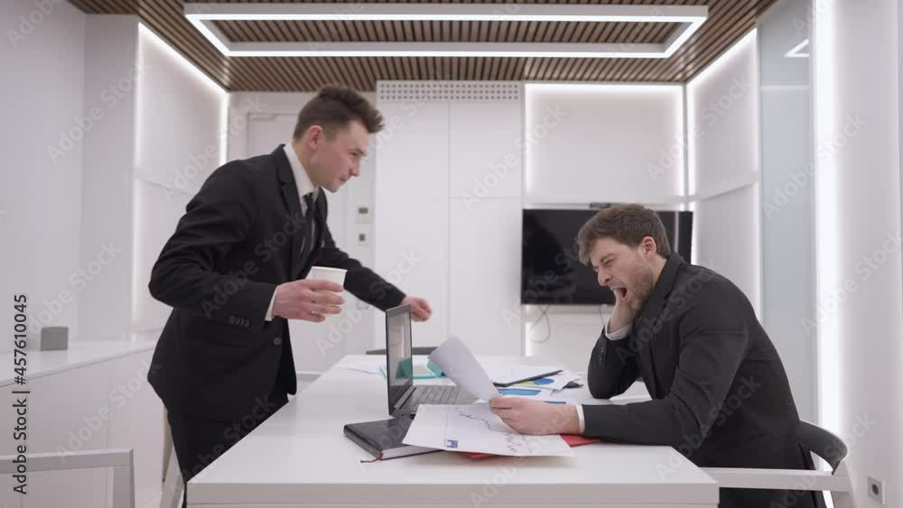 Caucasian man bringing coffee for overworked colleague sitting at table with laptop thinking. Side view portrait of positive coworkers supporting each other in office. Slow motion Caucasian man bringing coffee for overworked colleague sitting at table with laptop thinking. Side view portrait of positive coworkers supporting each other in office. Slow motion