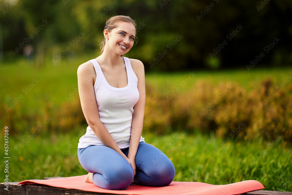 Smiling woman sits on exercising mat during yoga in city garden. Smiling woman sits on exercising mat during yoga in city garden.