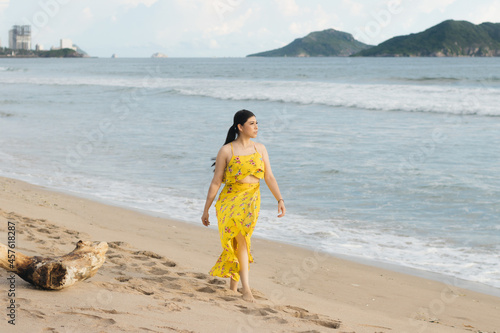 Young adult woman wearing a long yellow dress walking at the beach in the sunset
