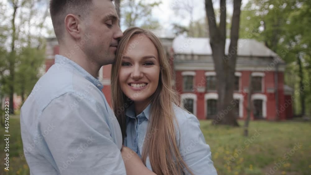 A beautiful young couple stands in the park against the background of a red building and are hugging looking at the camera. Slow motion A beautiful young couple stands in the park against the background of a red building and are hugging looking at the camera. Slow motion