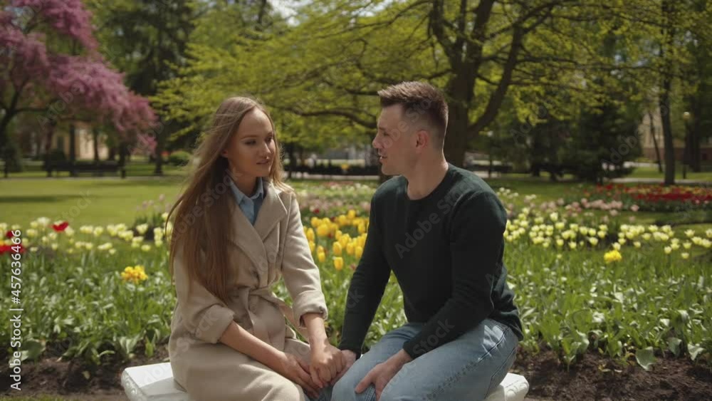 A beautiful young couple is talking and laughing while sitting on a bench against the backdrop of a blooming park. Slow motion A beautiful young couple is talking and laughing while sitting on a bench against the backdrop of a blooming park. Slow motion