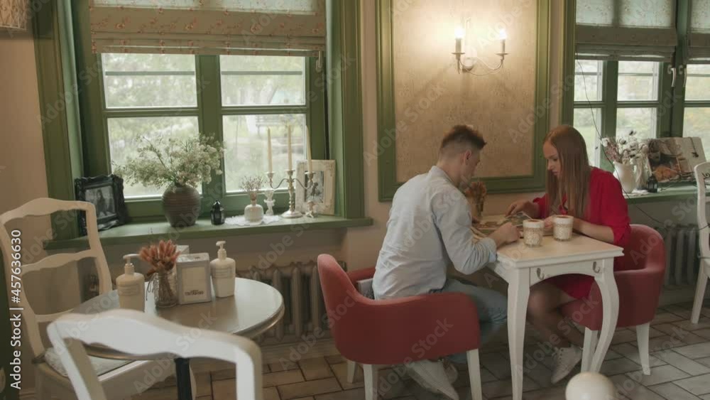 A young couple in love is sitting at a table in a cafe with a vintage interior and studying the menu. Side view. Slow motion A young couple in love is sitting at a table in a cafe with a vintage interior and studying the menu. Side view. Slow motion
