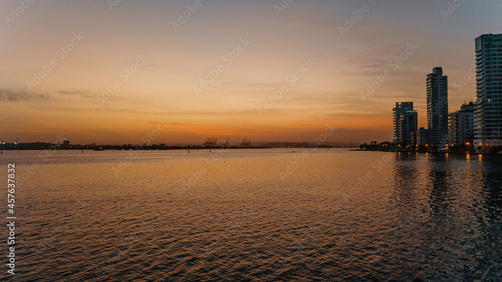 Atardecer o amanecer sobre la bahía con cielo naranja y amarillo con edificios, grúas y Sociedad Portuaria de fondo. Cartagena de Indias, Colombia