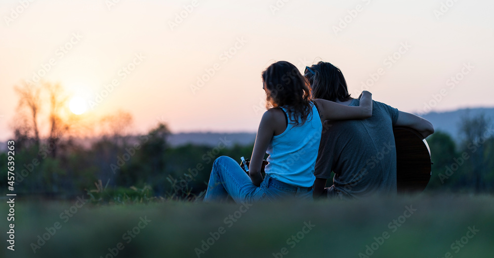 Couple sits on a hill and their boyfriend holds a guitar singing at sunset during a camping holiday. 