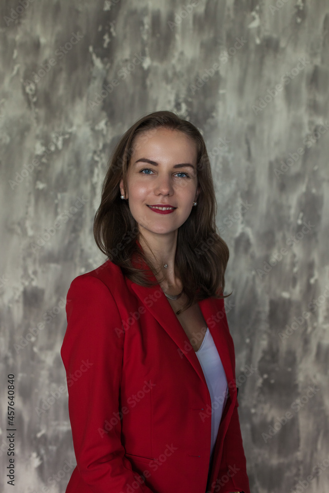 Portrait of a beautiful young business woman with brown hair in a red jacket. Side view of happy smiling girl with red lips on gray background with copy space. The concept of business and success. Portrait of a beautiful young business woman with brown hair in a red jacket. Side view of happy smiling girl with red lips on gray background with copy space. The concept of business and success.