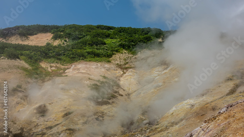 The soil on the hillside is covered with sulfur deposits. Steam and smoke rise from the fumaroles into the blue sky. Green vegetation on the top of the hill. Kamchatka