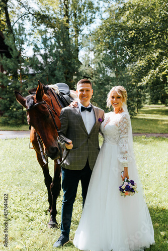 Happy beautiful young bride and groom hugging next to horse and looking at camera, married heterosexual couple in wedding suits in countryside. Wedding ceremony at ranch outdoors during the rain