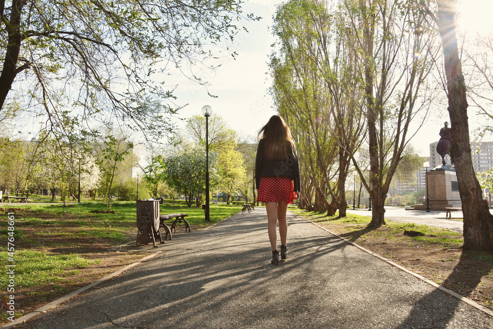 Full body back view of young beautiful slender woman walking on even ashalt road in park immersed in blooming trees in spring 