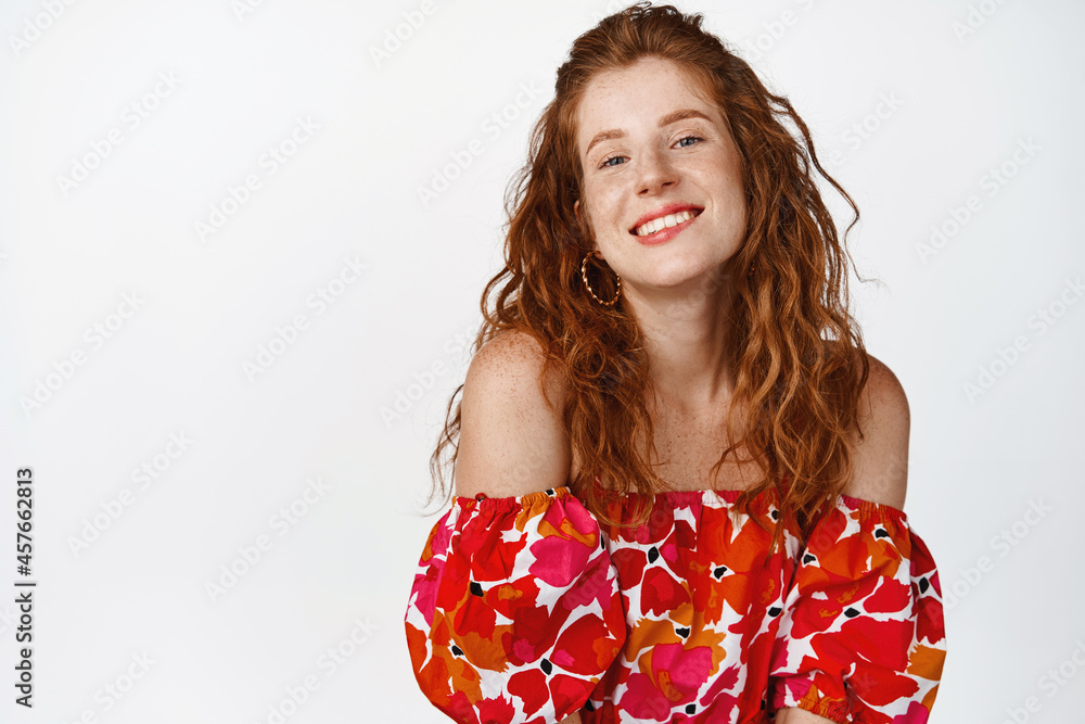 Coquettish redhead girl biting lip and looking flirty at camera, posing in stylish floral dress, standing against white background Coquettish redhead girl biting lip and looking flirty at camera, posing in stylish floral dress, standing against white background