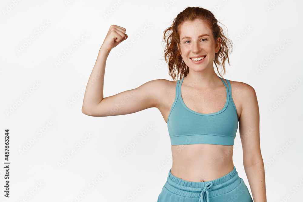 Active and healthy redhead sportswoman flexing biceps and smiling. Fitness girl workout, showing muscles on her arm, wearing sportsbra, standing over white background Active and healthy redhead sportswoman flexing biceps and smiling. Fitness girl workout, showing muscles on her arm, wearing sportsbra, standing over white background