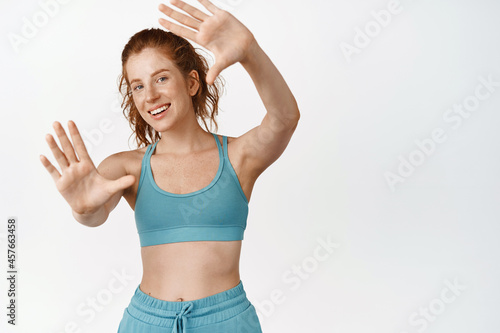 Beautiful redhead girl with fit healthy body, doing workout in gym, stretching out arms and smiling at camera, standing against white background