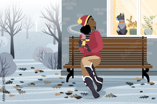 An African American girl sits on a bench in front of the house and looks at the cat in the window on a winter day. The girl is holding hot coffee in her hands.