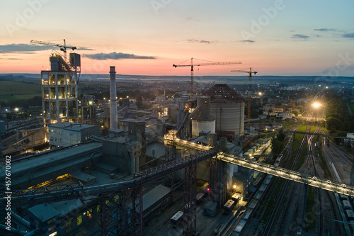 Aerial view of cement factory with high concrete plant structure and tower cranes at industrial production area at night. Manufacture and global industry concept.