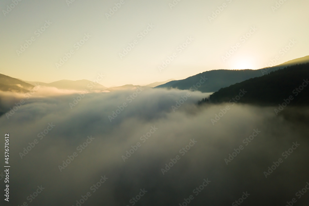 Aerial view of vibrant sunrise over white dense fog with distant dark silhouettes of mountain hills on horizon.