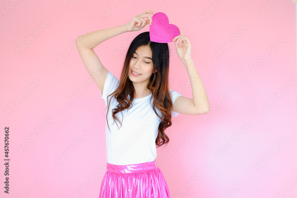 Portrait beautiful asian teen girl wearing white T-shirt and pink skirt on pink background, happy valentine day in love concept, model holding red heart sign in hand Portrait beautiful asian teen girl wearing white T-shirt and pink skirt on pink background, happy valentine day in love concept, model holding red heart sign in hand