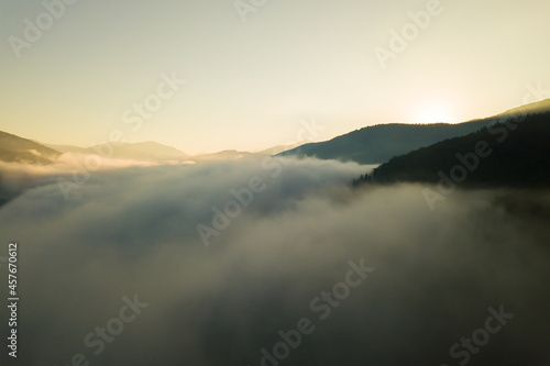 Aerial view of vibrant sunrise over white dense fog with distant dark silhouettes of mountain hills on horizon.