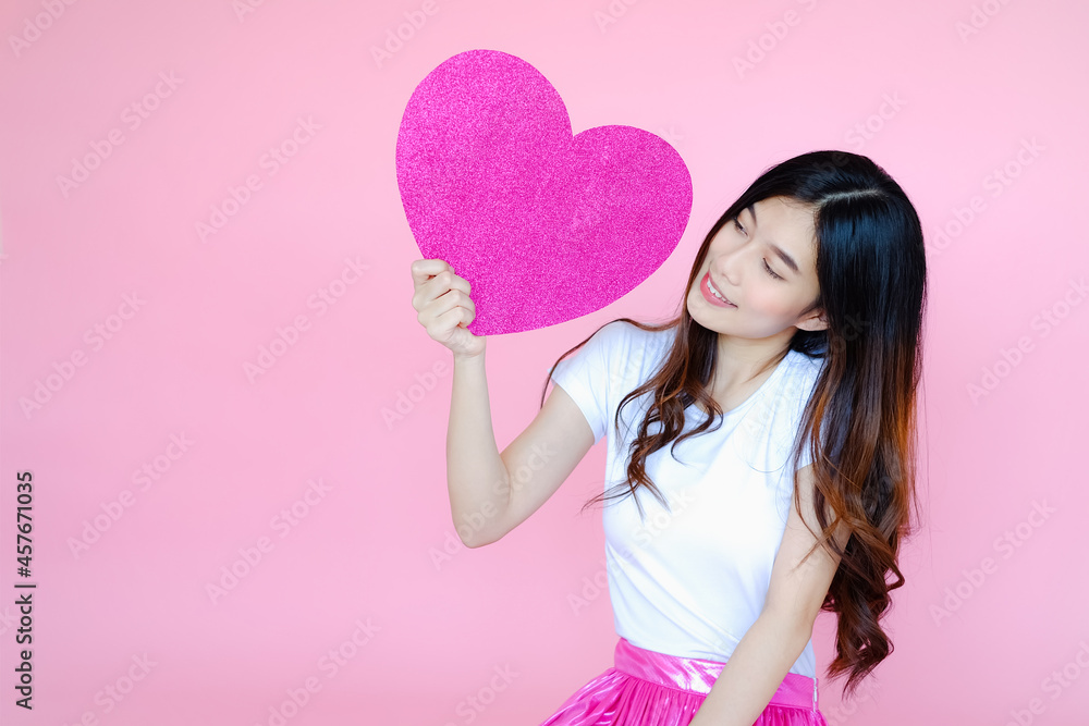 Portrait beautiful asian teen girl wearing white T-shirt and pink skirt on pink background, happy valentine day in love concept, model holding red heart sign in hand Portrait beautiful asian teen girl wearing white T-shirt and pink skirt on pink background, happy valentine day in love concept, model holding red heart sign in hand