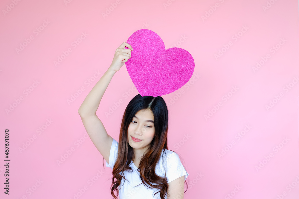 Portrait beautiful asian teen girl wearing white T-shirt and pink skirt on pink background, happy valentine day in love concept, model holding red heart sign in hand Portrait beautiful asian teen girl wearing white T-shirt and pink skirt on pink background, happy valentine day in love concept, model holding red heart sign in hand
