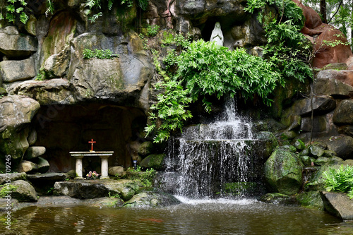 Fényképezés Statue of Our lady of grace virgin Mary view with natural background in Waterfall flows down from the rock cave at Thailand