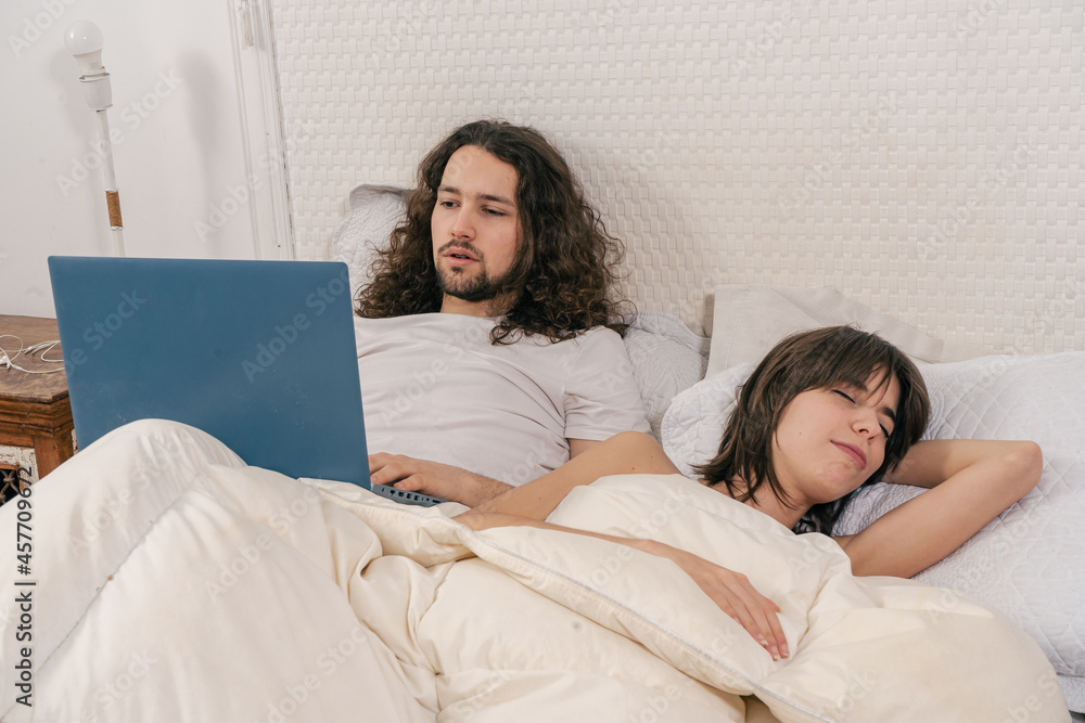 Couple in bed. The man entertaining with the laptop while the woman sleeps. Couple in bed. The man entertaining with the laptop while the woman sleeps.