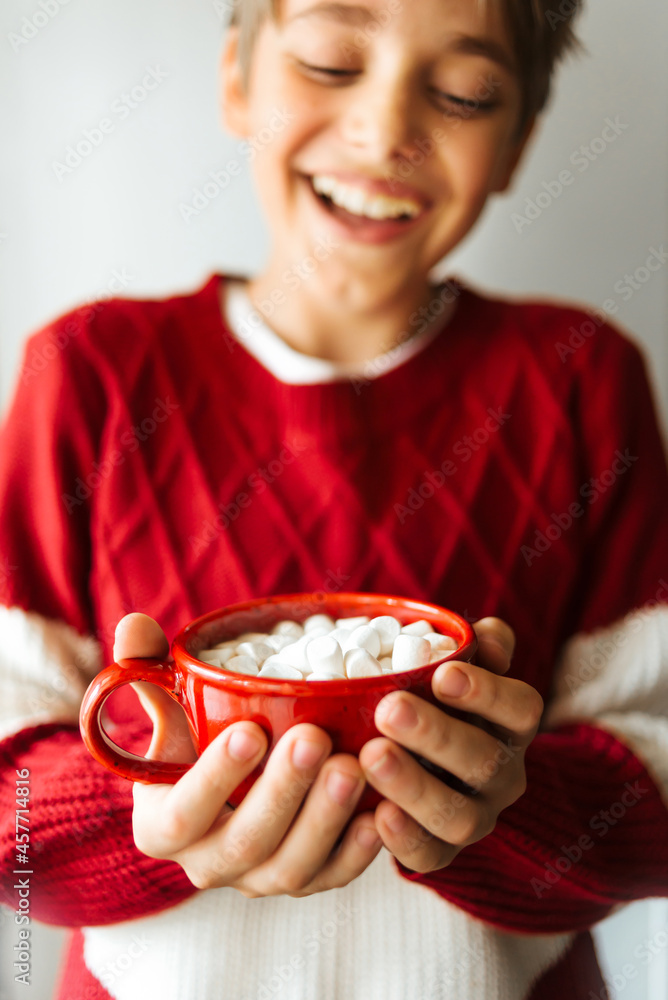 Happy smiling teenager boy holding a red cup with cocoa and marshmallows. Soft selective focus.