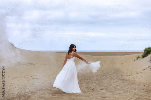 Junge schöne Frau im Hochzeitskleid am Strand mit einer Rauchfackel und weißen Rauch
