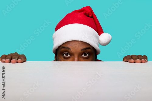 African-American man in a Santa Claus hat with expressive eyes looks intently at the camera looking out from under a white table on a turquoise background © Aleksandr