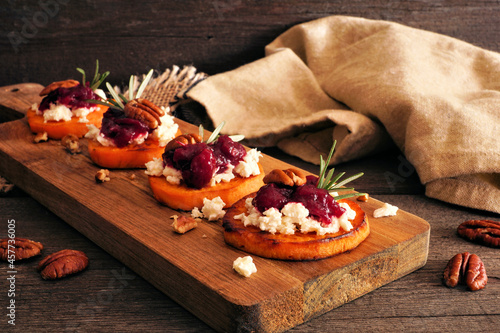 Autumn sweet potato crostini appetizers with cheese, cranberries and pecans on a wood platter. Table scene with a dark wood background.